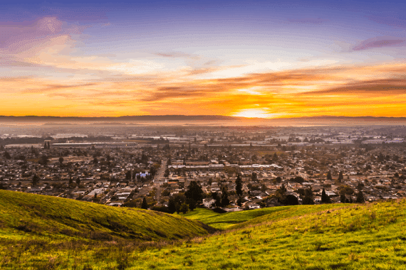 Sunset view of residential and industrial areas in East San Francisco Bay Area; green hills visible in the foreground; Hayward, California