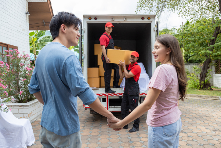 A young couple checks with two movers unloading boxes and furniture from a pickup truck.