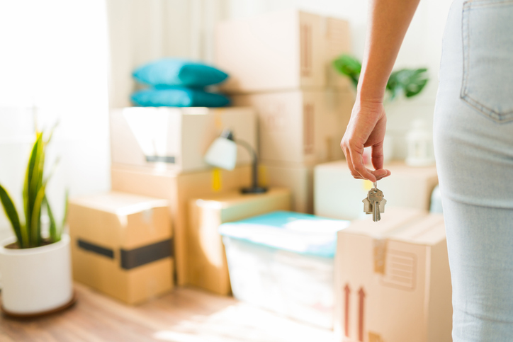 A young woman standing in her new living room, holding up a set of house keys, surrounded by stacked cardboard moving boxes, symbolizing the excitement and transition of moving to a new place.