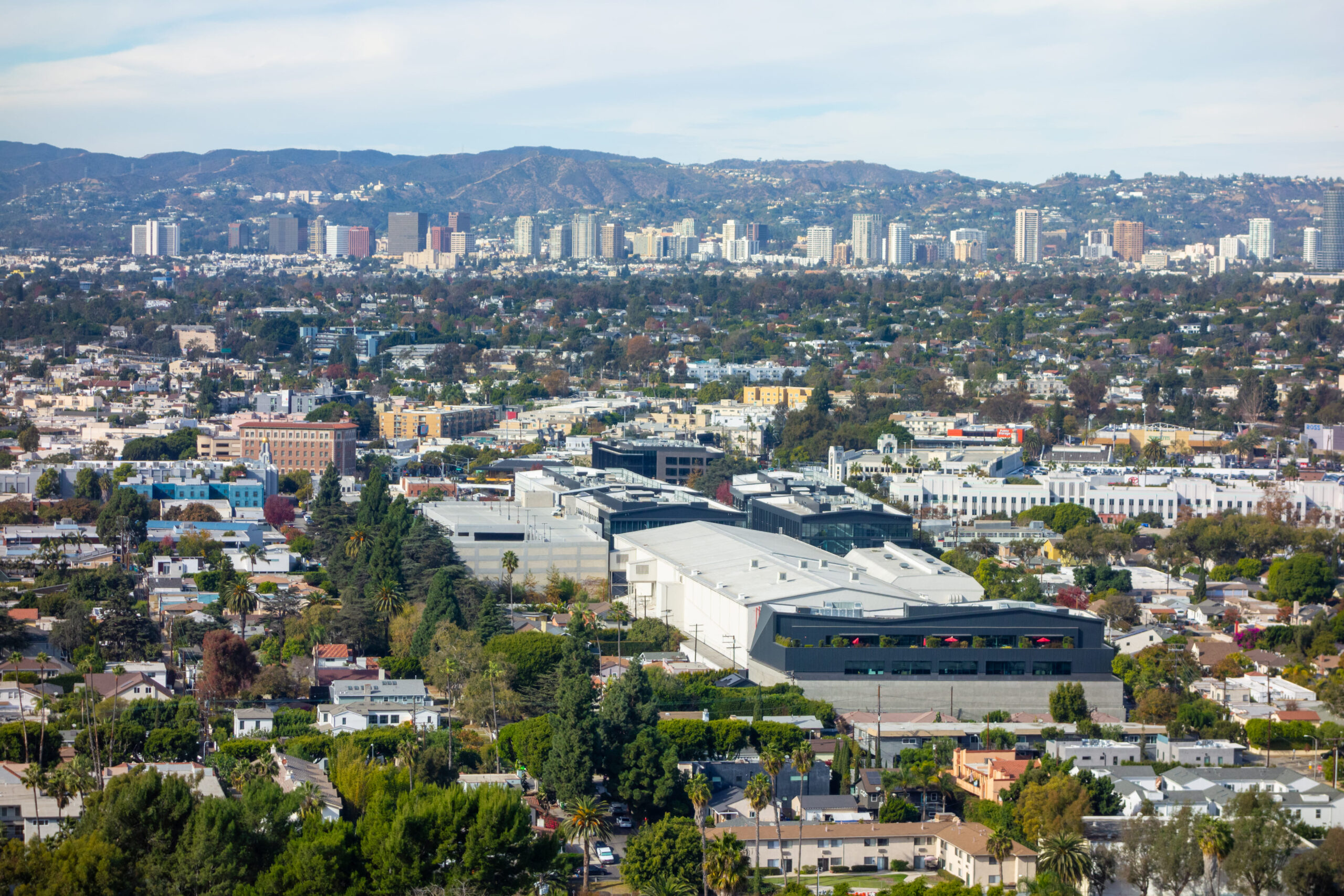A high-angle panoramic view captures the sprawling urban landscape of Culver City and Los Angeles, showing a mix of residential and commercial buildings nestled among green trees, with distant hills and mountains defining the background under a clear, blue sky.