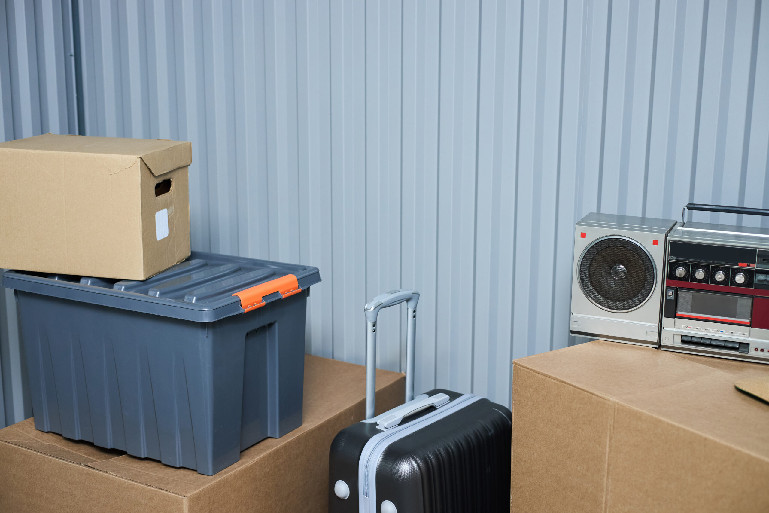 Stacked cardboard boxes, plastic storage container, hard shell suitcase with extended handle, vintage radio and cassette player arranged against corrugated metal wall in storage unit.