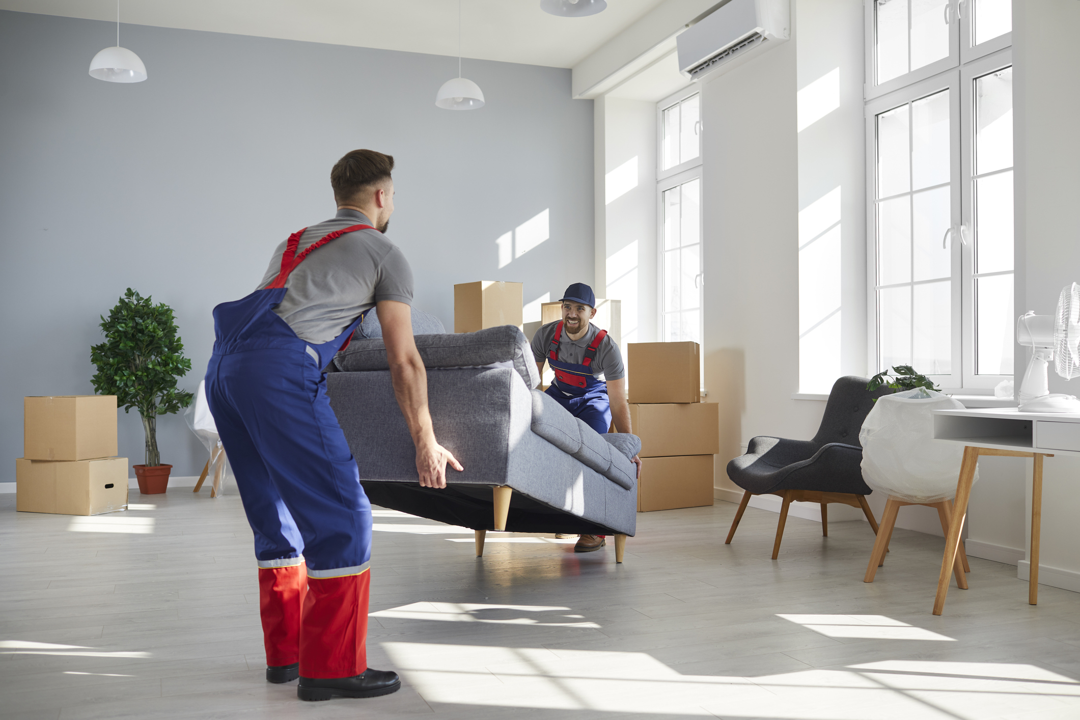 Two moving professionals lift up a heavy couch in a large living room with boxes and other furniture.